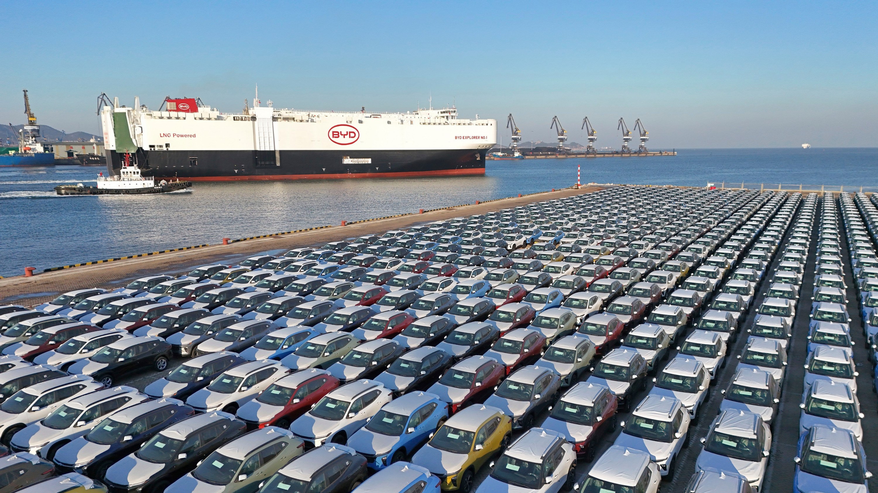 Thousands of cars are shown on a car carrier on a seaport, with a BYD freight boat in the background.