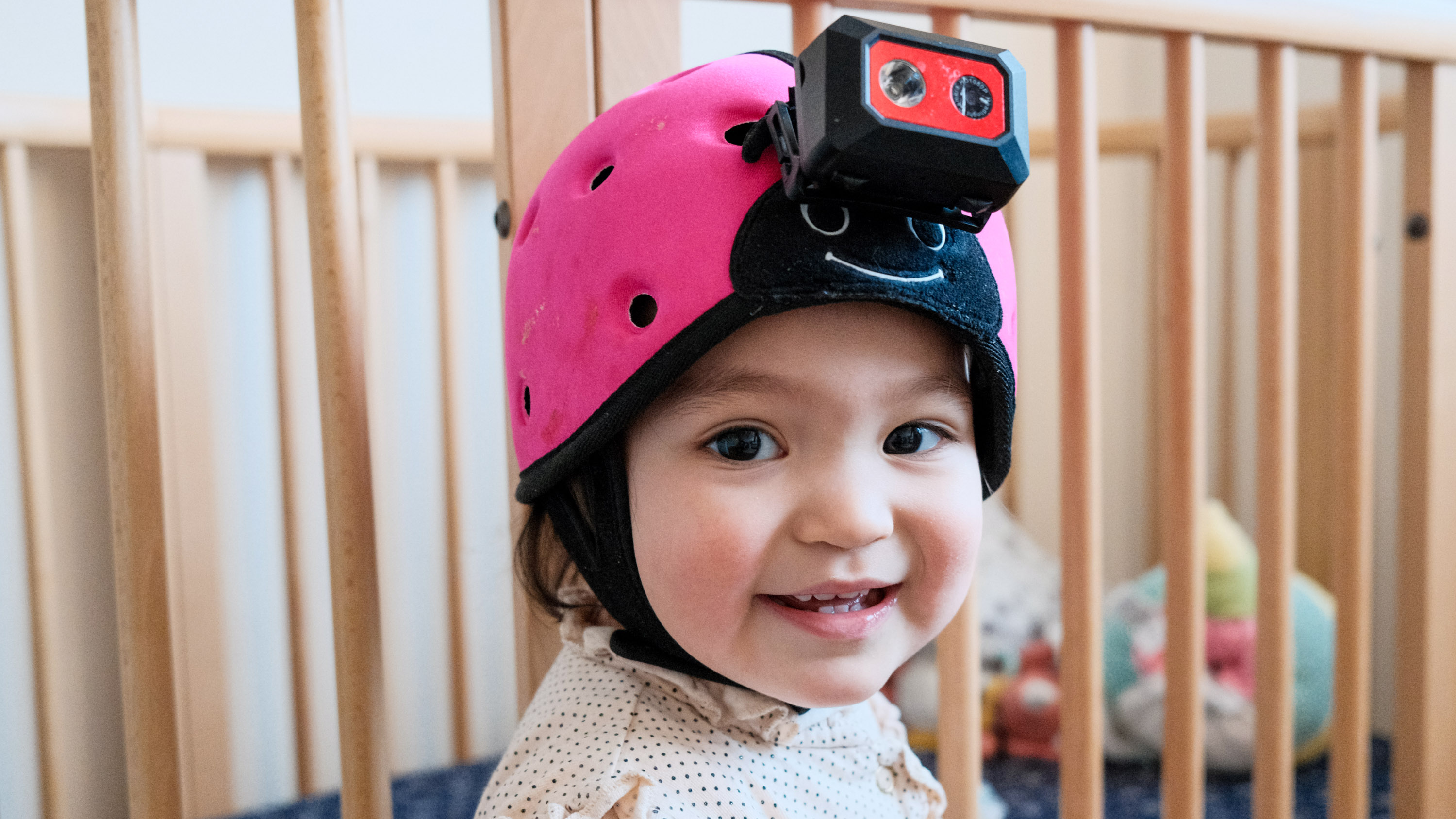 closeup of a smiling baby wearing a helmet camera with the bars of a crib in the background